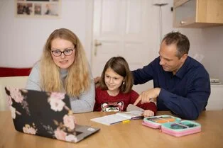 eine Frau, ein Mann und dazwischen ein Mädchen sitzen an einem Tisch. Die Frau hat einen aufgeklappten Laptop vor sich, das Kind Schulsachen. Die Erwachsenen scheinen ihr etwas zu erklären.